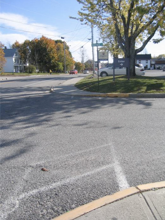 Image shows a faded crosswalk connecting two curbs with a tree and parking lot in the background. In the far distance, there is a person walking. 
